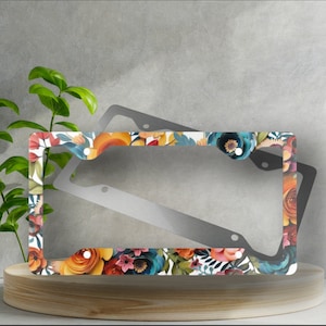 Puede incluir: Un marco de matrícula de metal con un diseño floral. El marco presenta un fondo blanco con flores y hojas coloridas. El marco está hecho de metal duradero y está diseñado para adaptarse a placas de matrícula estándar.
