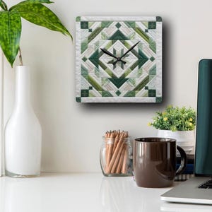 Op de afbeelding: Een vierkante wandklok met een groen en wit patchwork ontwerp. De wijzerplaat van de klok heeft zwarte wijzers en een witte achtergrond.