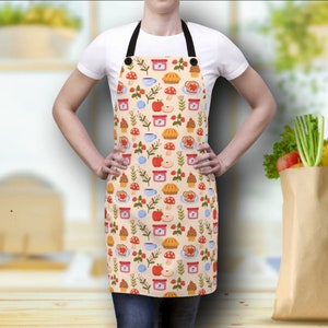Könnte beinhalten: Eine cremefarbene Küchenschürze mit einem bunten Muster aus Äpfeln, Kuchen, Kaffeetassen und Marmeladengläsern. Die Schürze hat verstellbare Träger und eine Tasche.