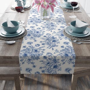 May include: A white table runner with a blue floral pattern. The runner is laid out on a wooden table with blue and white dishes and glasses.