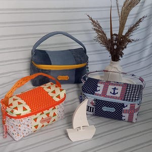 Könnte beinhalten: Drei handgefertigte Stofftaschen mit Griffen. Eine ist aus Jeansstoff mit einem gelben Reißverschluss, eine andere hat ein Wassermuster und die dritte zeigt ein Ankerdesign. Vor den Taschen steht eine kleine weiße Segelbootfigur.