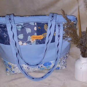 Könnte beinhalten: Handgefertigte Tragetasche mit hellblauem Grund und floral gemusterten Einsätzen in Blau- und Weißtönen. Die Tasche hat gestickte Griffe und ein kleines "Handmade"-Etikett. Im Hintergrund eine weiße Vase mit Trockenblumen.