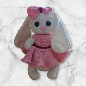 Peut inclure: Poupée lapin au crochet faite à la main, corps blanc, longues oreilles et nœud rose. Le lapin porte une robe rose avec une ceinture marron foncé. Yeux en boutons noirs et nez rose.