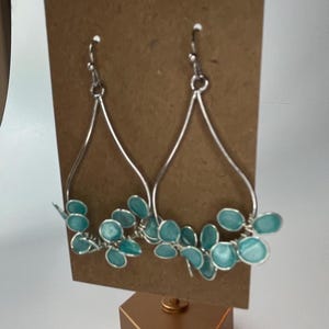 May include: A pair of silver dangle earrings with teal blue leaf-shaped charms. The earrings are displayed on a gold stand.