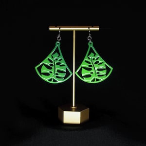 Pode incluir: Um par de brincos verdes em forma de folha com um design recortado, pendurados em um suporte dourado sobre um fundo preto.