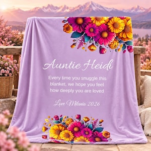 May include: A light purple blanket featuring a floral arrangement of yellow, orange, and magenta flowers at the top and bottom. The text reads: Auntie Heidi, Every time you snuggle this blanket, we hope you feel how deeply you are loved, Love Milania 2026.