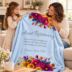 May include: A light blue fleece blanket featuring a floral border of pink, purple, and yellow flowers. The text reads: Aunt Rosemary, Every time you snuggle this blanket, we hope you feel how deeply you are loved, Christina, Nicole, Peter.