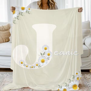 May include: Cream-colored blanket featuring a large white letter "J" adorned with daisy flowers. The blanket has a floral design with white daisies and yellow centers, and the word "eanie" is printed on the blanket.