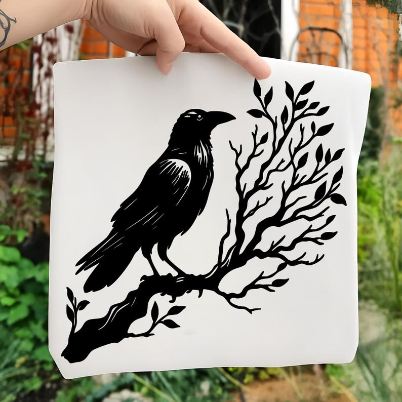 May include: A white tote bag featuring a black illustration of a raven perched on a tree branch. The raven is detailed with visible feathers and a sharp beak. The tree branch has leaves and a textured appearance.