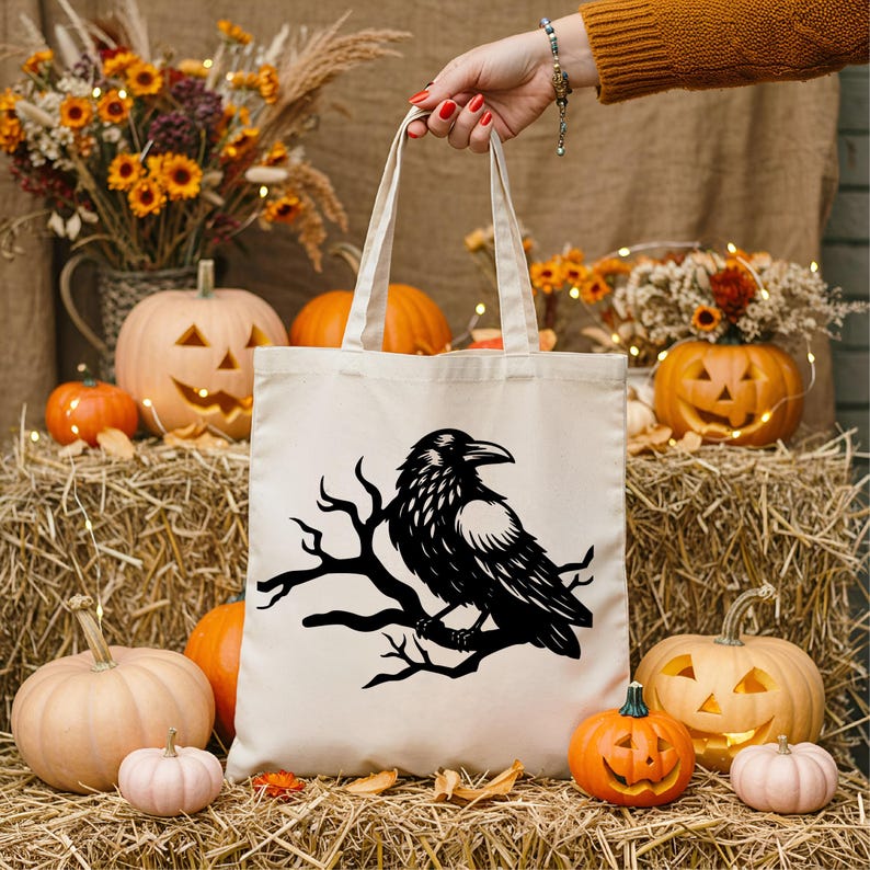 May include: A cream-colored tote bag featuring a black raven perched on a branch. The bag is held by a person and is surrounded by pumpkins, gourds, and fall foliage. The scene evokes a Halloween or autumn theme.