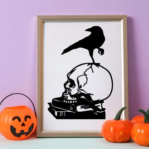 May include: A framed black and white print featuring a skull with a raven perched on top. The skull rests on a stack of books. A jack-o'-lantern bucket and pumpkins are in the foreground, creating a Halloween theme.