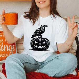 May include: White t-shirt featuring a black graphic of a raven perched on a carved pumpkin. The pumpkin has a jack-o'-lantern face. The shirt is a casual style, perfect for Halloween or fall. The raven and pumpkin design is a classic Halloween motif.