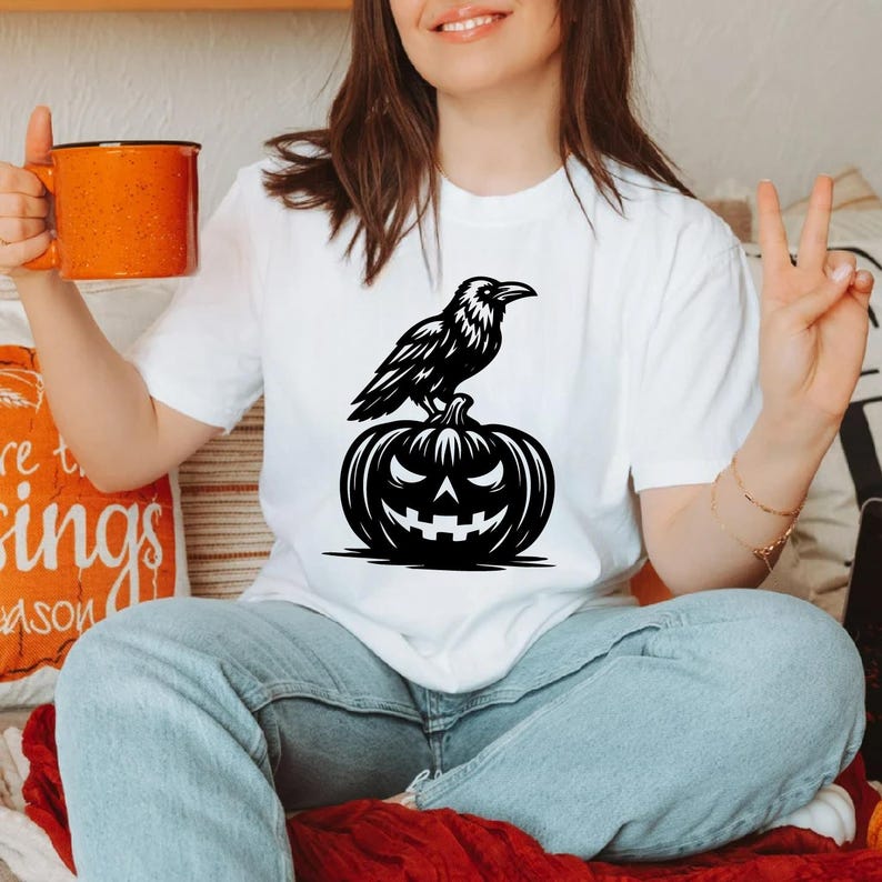 May include: White t-shirt featuring a black graphic of a raven perched on a carved pumpkin. The pumpkin has a jack-o'-lantern face. The shirt is a casual style, perfect for Halloween or fall. The raven and pumpkin design is a classic Halloween motif.
