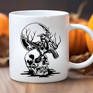 May include: White ceramic mug featuring a black graphic of a skull, a raven perched on a branch, and a full moon. The design is set against a blurred background with pumpkins, suggesting a Halloween theme.