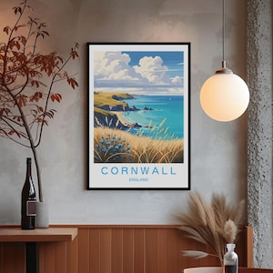 May include: A framed art print featuring a coastal landscape of Cornwall, England. The artwork depicts a vibrant scene with turquoise water, golden cliffs, and a blue sky with fluffy white clouds. The word "CORNWALL" is printed in white, with "ENGLAND" below it.