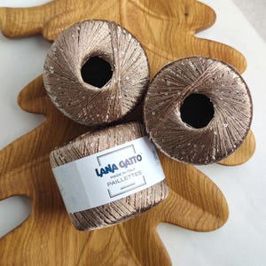 Può includere: Tre gomitoli di filato color bronzo metallizzato con piccole paillettes argentate integrate. Un gomitolo presenta un'etichetta di carta bianca con il testo Lana Gatto, Made in Italy e Paillettes stampato in nero, avvolto attorno al filo.