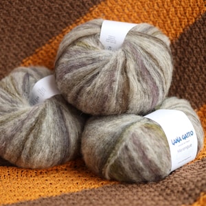 May include: Three round balls of Lana Gatto Meringue yarn featuring a variegated blend of soft grey, beige, and muted purple fibres. Each ball is wrapped with a white paper label displaying the brand name and product line in blue text.