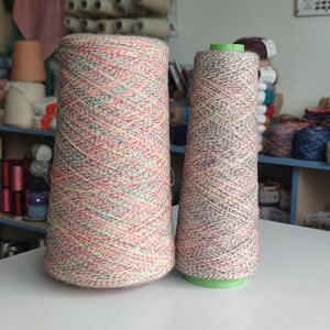 Pode incluir: Dois grandes cones de fio com um padrão salpicado multicolorido, apresentando fibras vermelhas, verdes, amarelas e azuis torcidas num fio de base claro. O fio está enrolado firmemente à volta dos cones de plástico centrais.