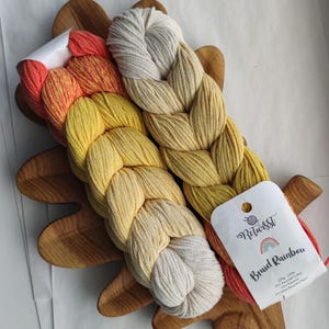 May include: A selection of braided yarn in yellow, orange, and cream hues, presented on a wooden leaf-shaped tray. The yarn is labelled "Braid Rainbow" with a rainbow graphic. The yarn is made of 70% recycled cotton, 25% polyester, and 5% other recycled fibres.