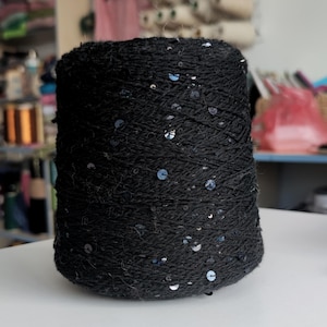 May include: A large cylindrical spool of black yarn featuring small, reflective silver sequins distributed throughout the fiber. The yarn has a textured, slightly fuzzy appearance, and the sequins catch the light, creating a subtle sparkle across the dark surface of the spool.