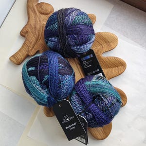 Peut inclure: Trois pelotes de laine multicolores dans des tons de violet profond, bleu roi et bleu sarcelle. Chaque pelote comporte une étiquette en papier noir avec le texte Schoppel Zauberball Crazy et 100g imprimé, indiquant le poids de la laine.