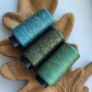 May include: Three cylindrical spools of metallic thread arranged diagonally. The top spool is teal, the middle is olive green, and the bottom is bright emerald green. Each spool features a shiny, reflective texture with a black plastic base.