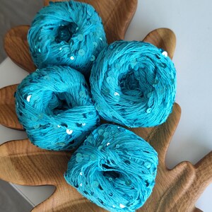 May include: Four round balls of bright turquoise yarn, each intricately woven with small, reflective circular sequins that catch the light. The yarn has a textured, fibrous appearance, and the sequins are distributed evenly throughout the material.