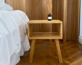 May include: A light brown wooden nightstand with a rectangular open shelf design and four tapered legs. A small dark glass bottle with a white label and several wooden reed diffuser sticks is placed on the top surface of the nightstand.