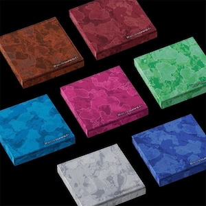 Op de afbeelding: Een assortiment vierkante dozen in verschillende kleuren, waaronder bruin, rood, groen, roze, blauw en grijs. Elke doos heeft een marmerpatroon en de tekst "the/incunabula". De dozen zijn geplaatst op een zwarte ondergrond.