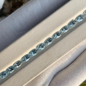 May include: A delicate bracelet featuring a series of oval-cut, light blue gemstones. The gemstones are set in a row, creating a sparkling and elegant accessory. The bracelet is displayed on a white surface, highlighting the color and clarity of the stones.