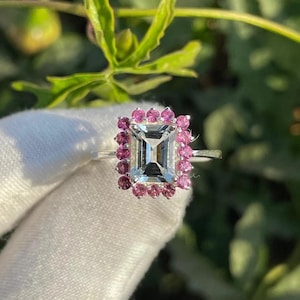 May include: A silver ring with a rectangular, clear gemstone at its centre, encircled by small, round, pink gemstones. The ring is set against a blurred green and white backdrop, accentuating the ring's design.