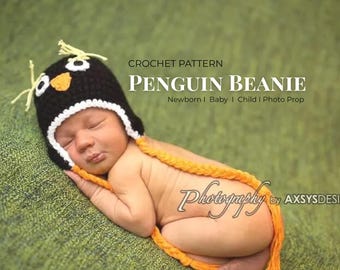 May include: A newborn baby wearing a black and white crochet penguin beanie with yellow accents. The beanie has a white trim and yellow yarn details. The baby is lying on a green surface, with a yellow yarn tail.