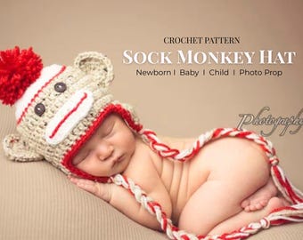 May include: A newborn baby wearing a crocheted sock monkey hat with a red pom-pom and a red and white striped trim. The baby is curled up on a neutral-colored surface, with a red and white rope-like trim around the body.