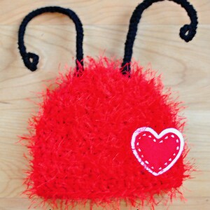 May include: A handmade red fuzzy hat with black antennae and a heart applique. The hat is made of a soft, textured material and features a white-stitched heart on the front. The text "monkey shine designs" is at the bottom.