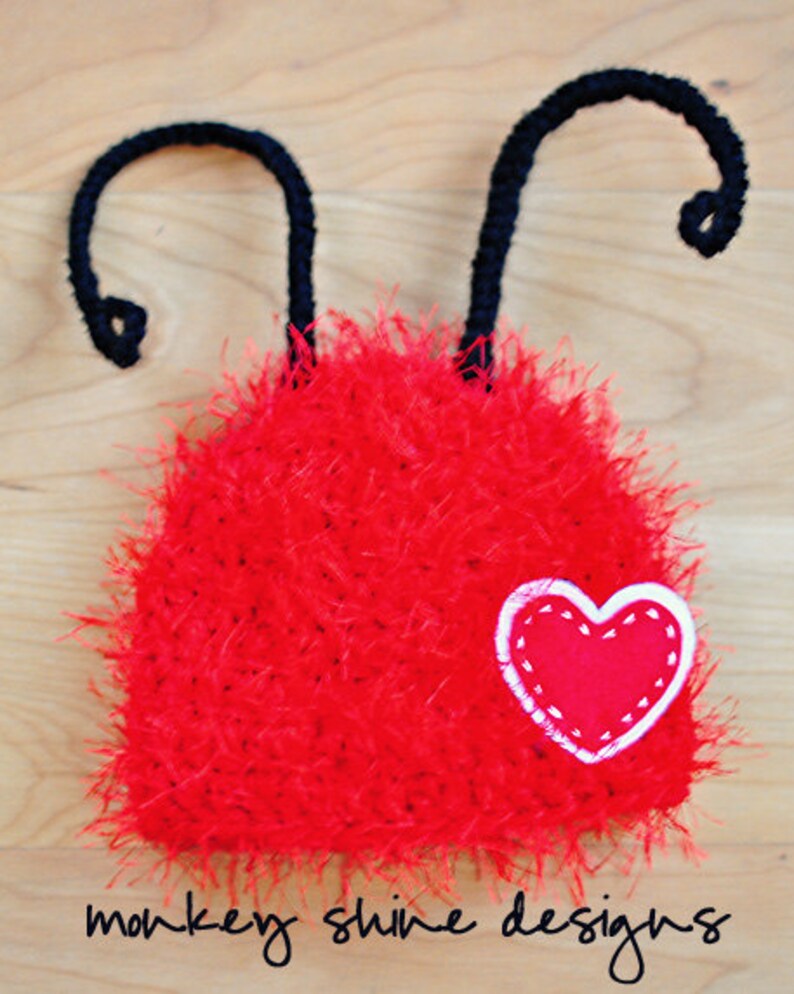 May include: A handmade red fuzzy hat with black antennae and a heart applique. The hat is made of a soft, textured material and features a white-stitched heart on the front. The text "monkey shine designs" is at the bottom.