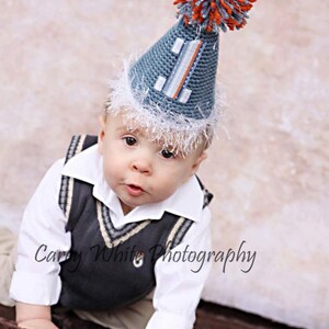Pode incluir: Um beb&ecirc; usando um chap&eacute;u de festa azul de malha com o n&uacute;mero "1" e um pompom fofo laranja e azul. O beb&ecirc; tamb&eacute;m est&aacute; usando uma camisa branca com gola e um colete cinza com acabamento branco.