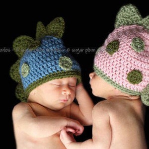 May include: Two baby hats in a dinosaur design. One hat is blue with green accents, and the other is pink with green accents. The hats have a textured, crocheted appearance and are adorned with circular green details.