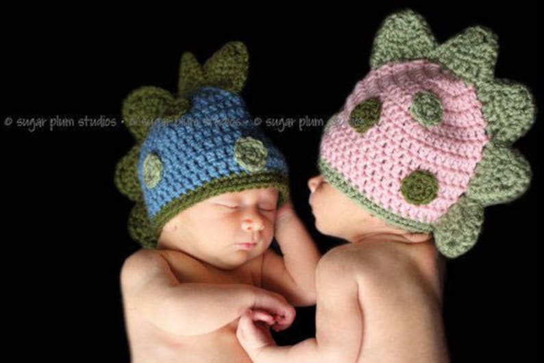 May include: Two baby hats in a dinosaur design. One hat is blue with green accents, and the other is pink with green accents. The hats have a textured, crocheted appearance and are adorned with circular green details.