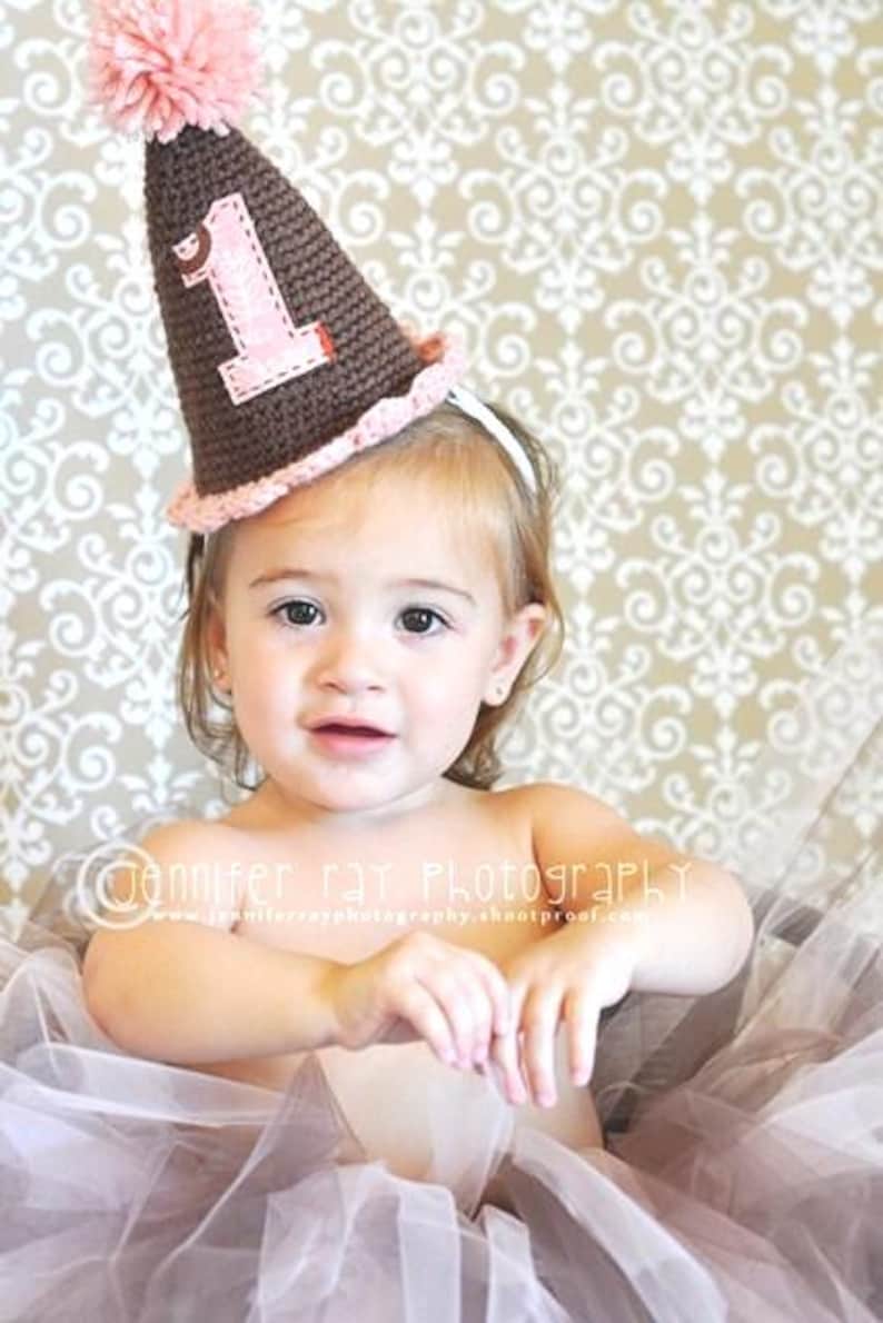 Pode incluir: Um chap&eacute;u de festa de anivers&aacute;rio de croch&ecirc; marrom e rosa com o n&uacute;mero "1" na frente. O chap&eacute;u tem um pompom rosa no topo e uma borda rosa. A crian&ccedil;a est&aacute; usando uma faixa de cabelo branca e um tutu rosa.