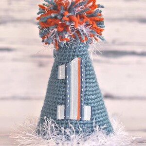 Pode incluir: Um chap&eacute;u de festa de croch&ecirc; azul feito &agrave; m&atilde;o com um aplique com o n&uacute;mero 1. O chap&eacute;u &eacute; coberto com um pompom de fio laranja, azul e branco. A base do chap&eacute;u tem franjas brancas. O texto "monkey shine designs" est&aacute; na parte inferior.