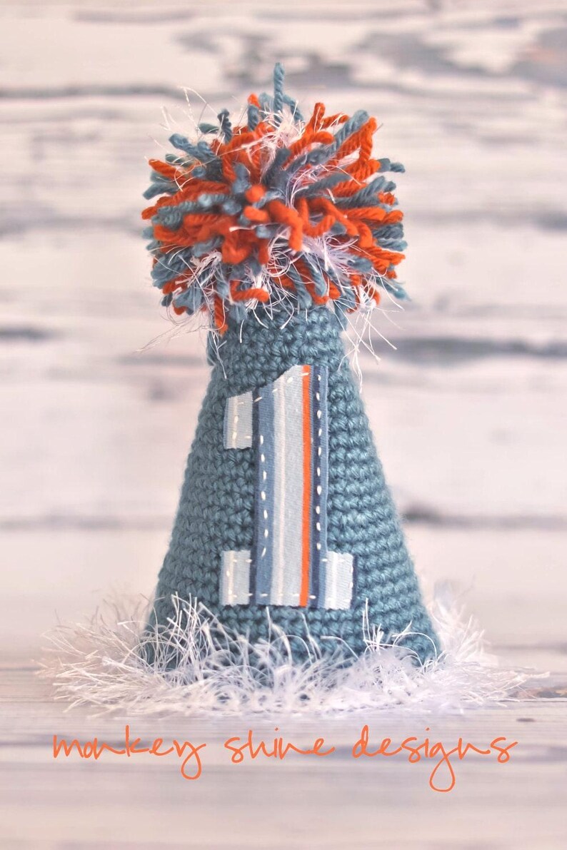 Pode incluir: Um chap&eacute;u de festa de croch&ecirc; azul feito &agrave; m&atilde;o com um aplique com o n&uacute;mero 1. O chap&eacute;u &eacute; coberto com um pompom de fio laranja, azul e branco. A base do chap&eacute;u tem franjas brancas. O texto "monkey shine designs" est&aacute; na parte inferior.