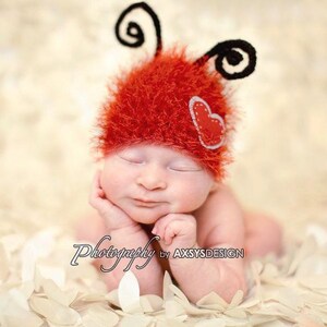 May include: A newborn baby wearing a red, fuzzy ladybug hat with black antennae and a heart applique. The baby is lying on a bed of white petals, with eyes closed. The image is labeled "Photography by AXSYS DESIGN".