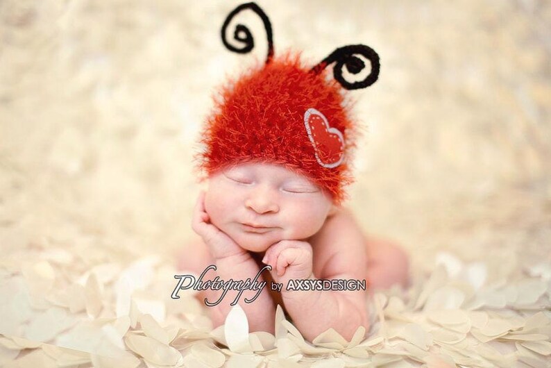 May include: A newborn baby wearing a red, fuzzy ladybug hat with black antennae and a heart applique. The baby is lying on a bed of white petals, with eyes closed. The image is labeled "Photography by AXSYS DESIGN".