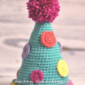 Pode incluir: Um chap&eacute;u de festa de croch&ecirc; turquesa feito &agrave; m&atilde;o com bolinhas coloridas e um grande pompom f&uacute;csia no topo. A base do chap&eacute;u tem uma borda de arco-&iacute;ris e as palavras "monkey shine designs" est&atilde;o escritas na lateral.
