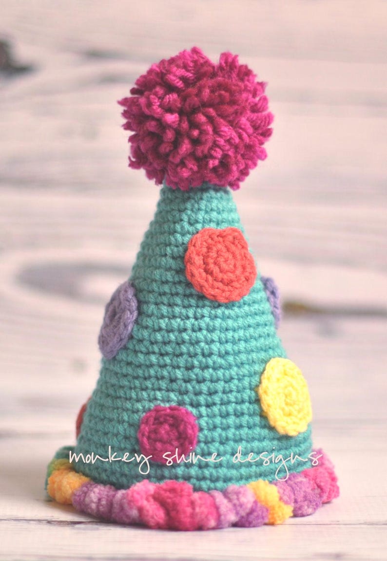 Pode incluir: Um chap&eacute;u de festa de croch&ecirc; turquesa feito &agrave; m&atilde;o com bolinhas coloridas e um grande pompom f&uacute;csia no topo. A base do chap&eacute;u tem uma borda de arco-&iacute;ris e as palavras "monkey shine designs" est&atilde;o escritas na lateral.
