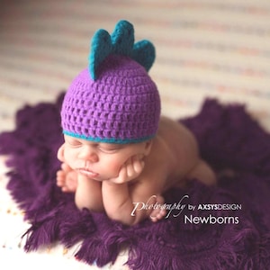 May include: A newborn baby wearing a purple crocheted hat with teal accents, resting on a matching purple blanket. The hat has a spiky design. The image is labeled "Newborns" and "Photography by AXSYSDESIGN".