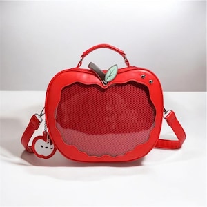Peut inclure: Un sac à main rouge en forme de pomme avec une poignée et une bandoulière amovible. Le sac comporte une fenêtre en filet et un petit charme de pomme. Le sac a une feuille verte et une tige marron.