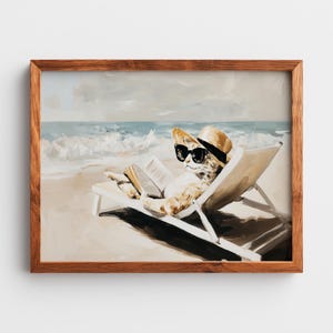 May include: A framed wall art print featuring a painting of a light-colored cat wearing a straw hat and black sunglasses while reclining on a white beach chair and reading an open book, set against a backdrop of a sandy beach and blue ocean waves.