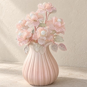 May include: A pale pink ceramic vase with a ribbed, scalloped rim containing a bouquet of translucent light pink glass flowers. Each flower features a central white pearl-like bead and is accompanied by delicate, pale green glass leaves.