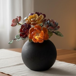 以下が含まれることがあります： 光沢のある色とりどりの陶器の花束が入った球形のマットブラックの花瓶。オレンジ、黄色、濃い赤、淡いピンクの色合いの花びらと緑の葉が特徴で、暗い丸いベースに対して鮮やかなコントラストを生み出しています。